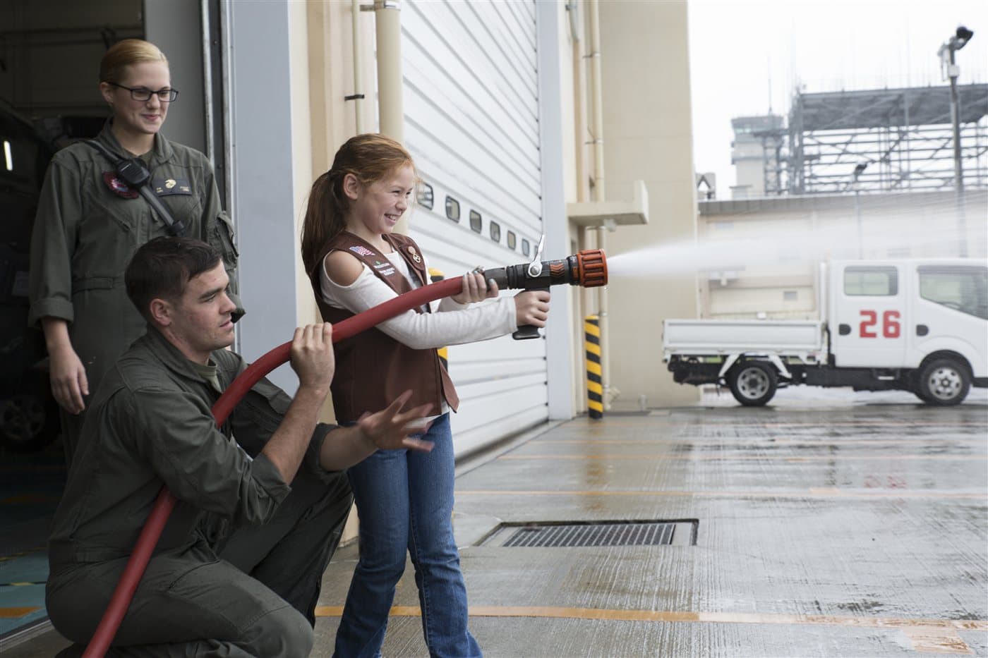 A smiling Brownie Girl Scout spraying a real fire hose with two firefighters helping her